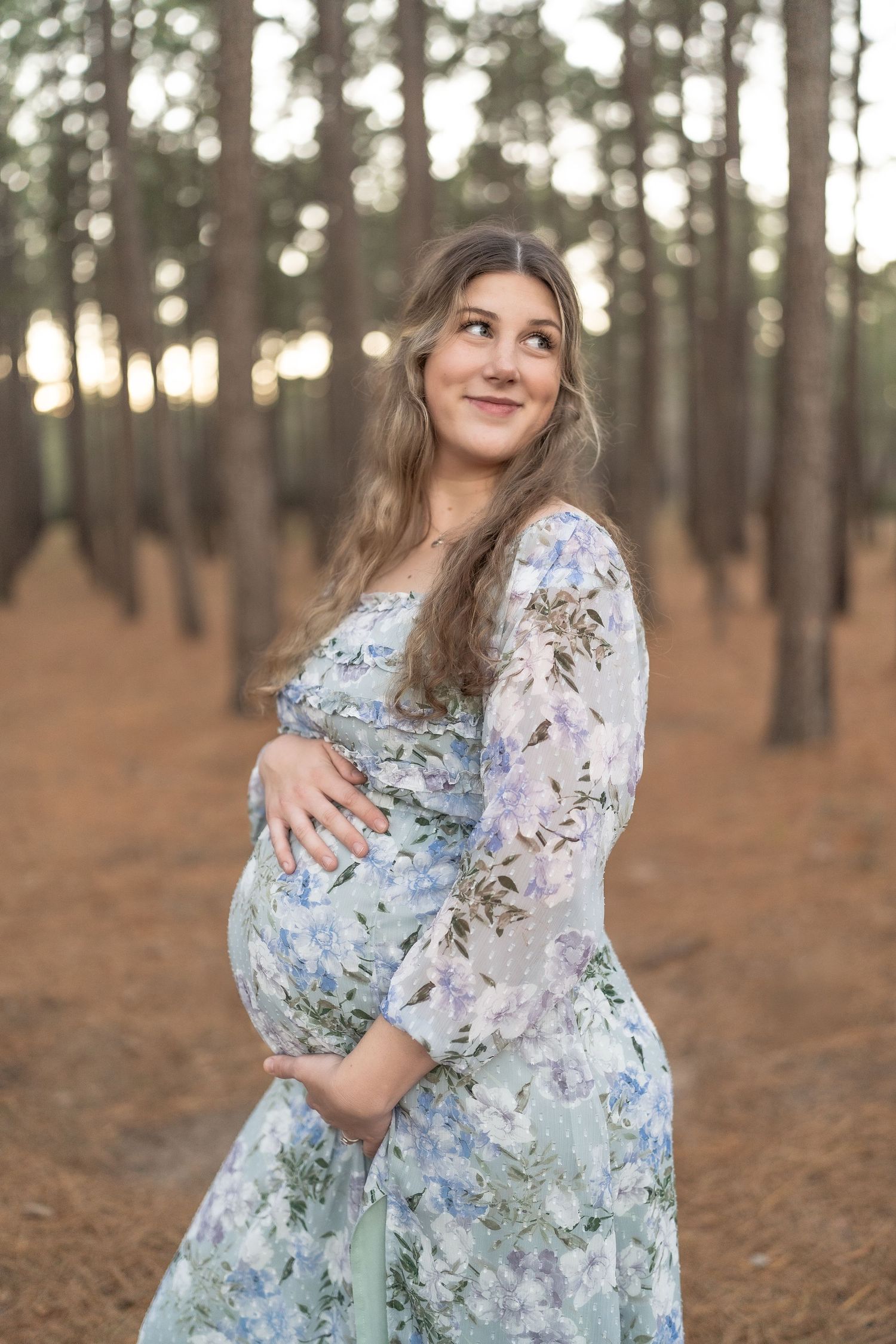 Pregnant woman in floral dress cradling baby bump in pine forest, smiling upward in golden light.