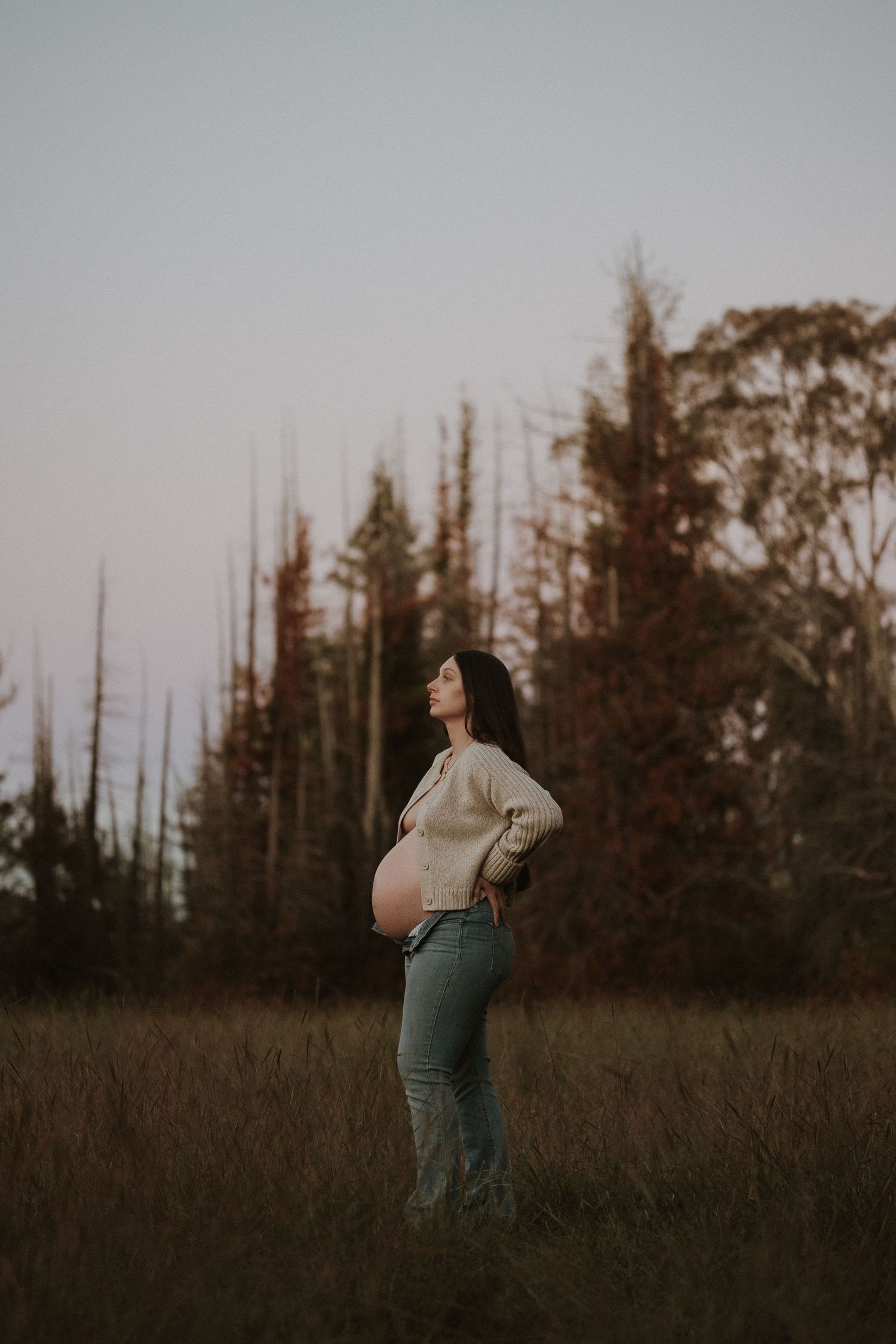 Pregnant woman in beige top and jeans standing in a field at dusk, cradling her baby bump with trees in background.