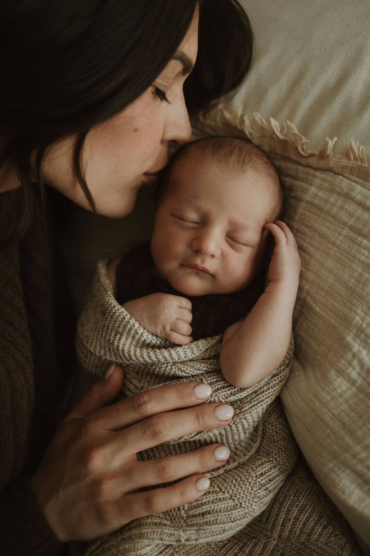 A mother tenderly kisses her sleeping newborn baby wrapped in a knit blanket during a newborn photography session.