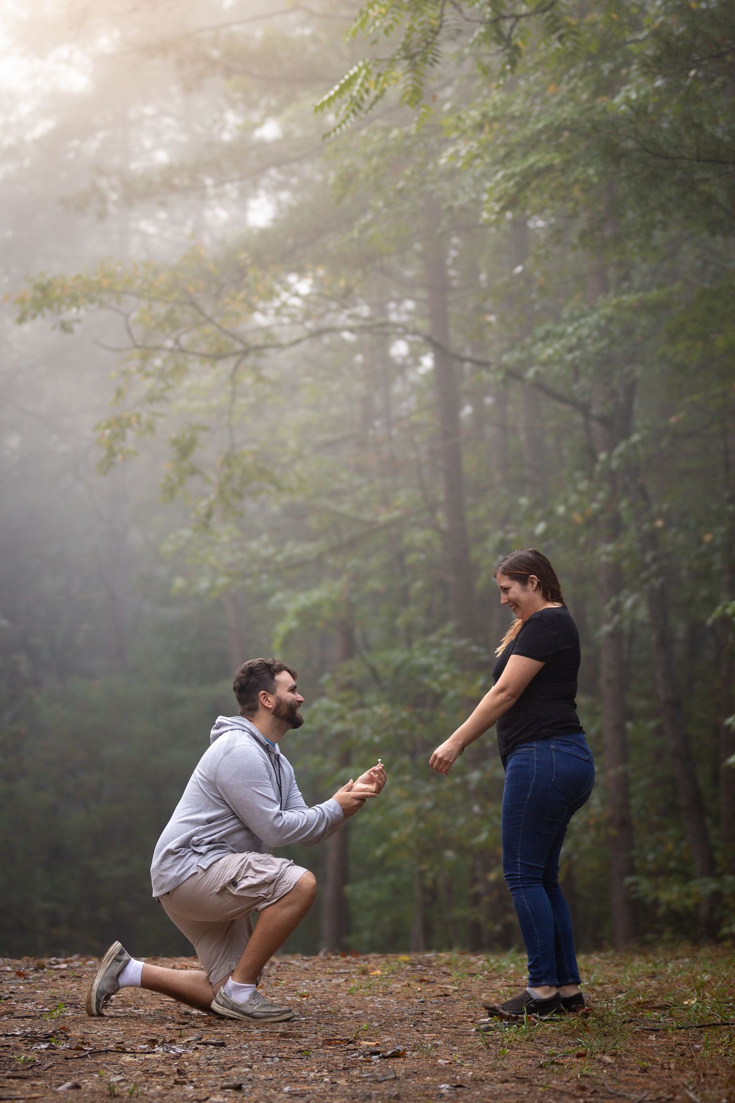 Man proposing on one knee to woman in misty forest, holding ring box in romantic outdoor marriage proposal scene.