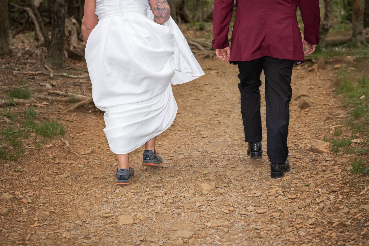 Bride in white dress and blue sneakers walks hand-in-hand with groom in burgundy suit on a woodland path.