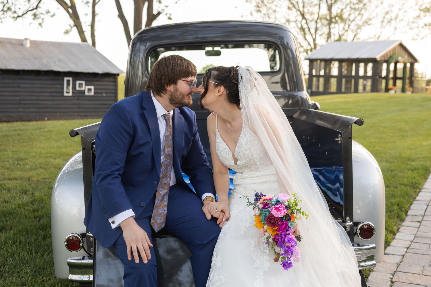 Newlywed couple sharing a romantic moment seated in the bed of a vintage truck, bride holding colorful bouquet.