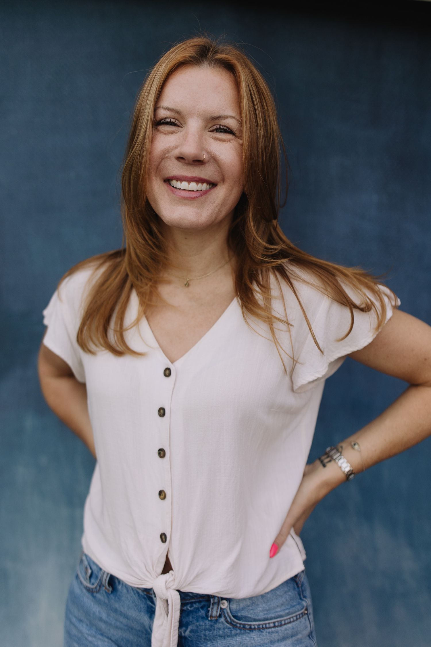 Smiling woman with auburn hair wearing a white button-up top and jeans, posing against a blue background.