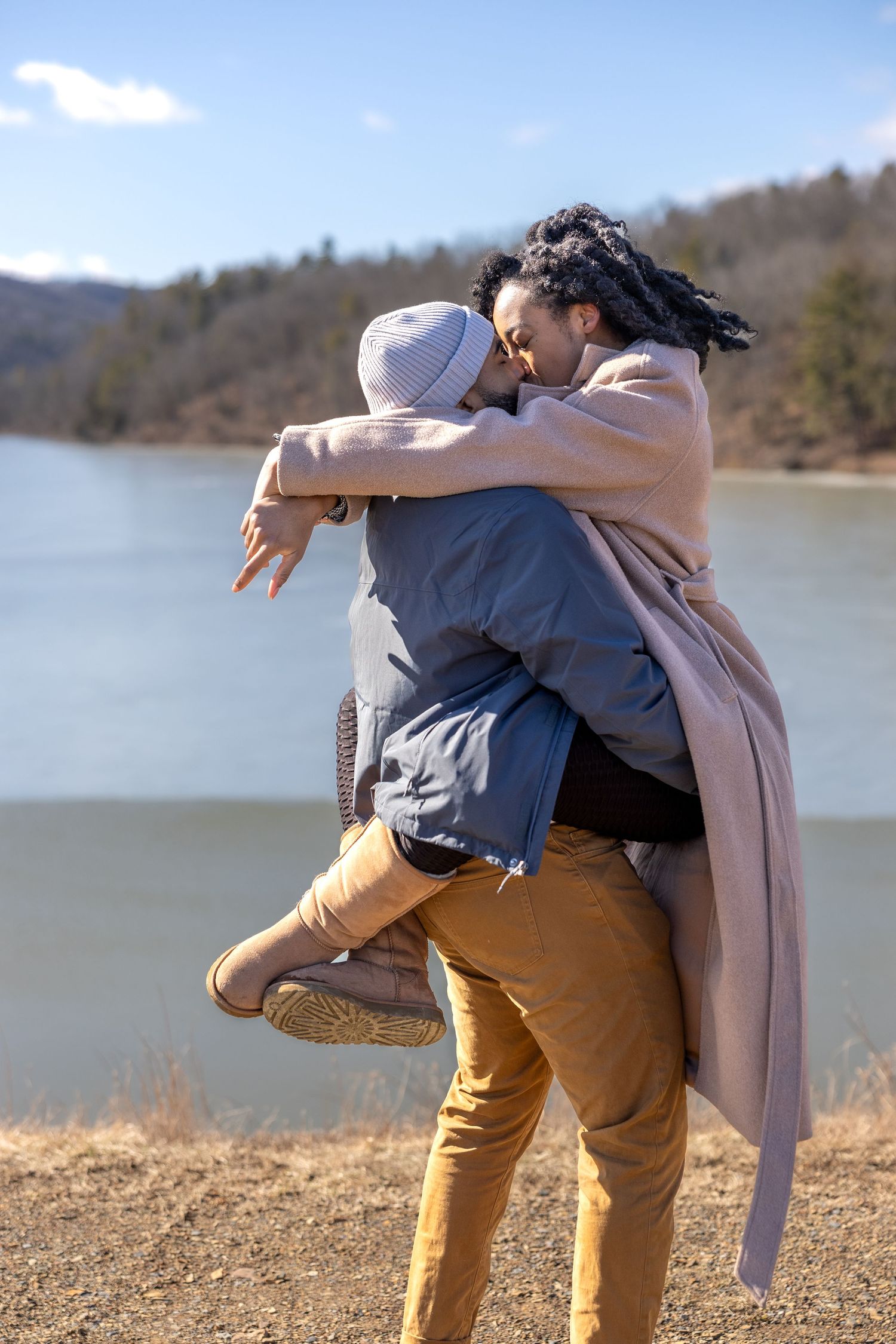 Couple embracing outdoors near a lake on a sunny winter day, man lifting woman in a romantic hug.