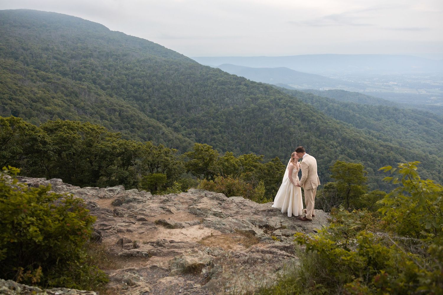 Bride and groom share a romantic kiss on a rocky mountain overlook surrounded by lush green forested hills.