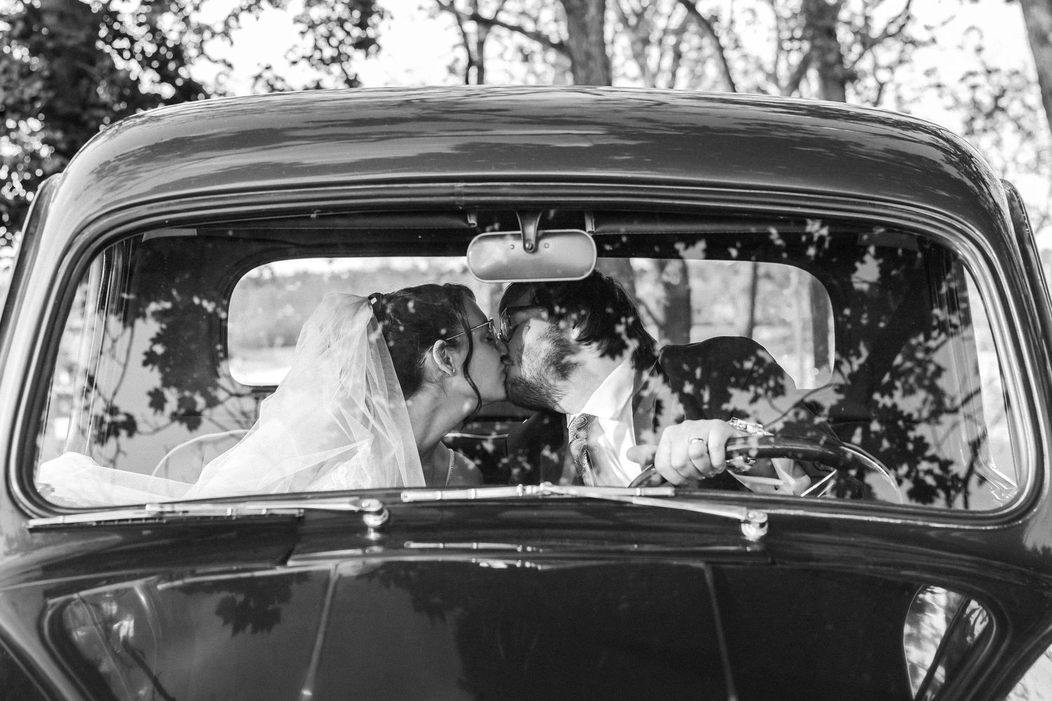 Black and white photo of a bride and groom sharing a kiss inside a vintage classic car on their wedding day.