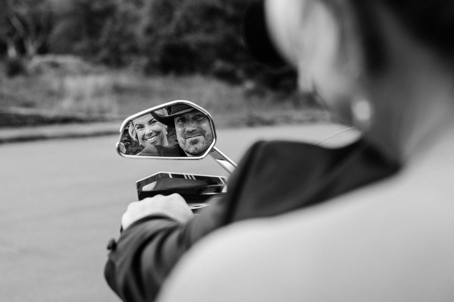 Black and white photo of a couple smiling, reflected in a motorcycle side mirror.