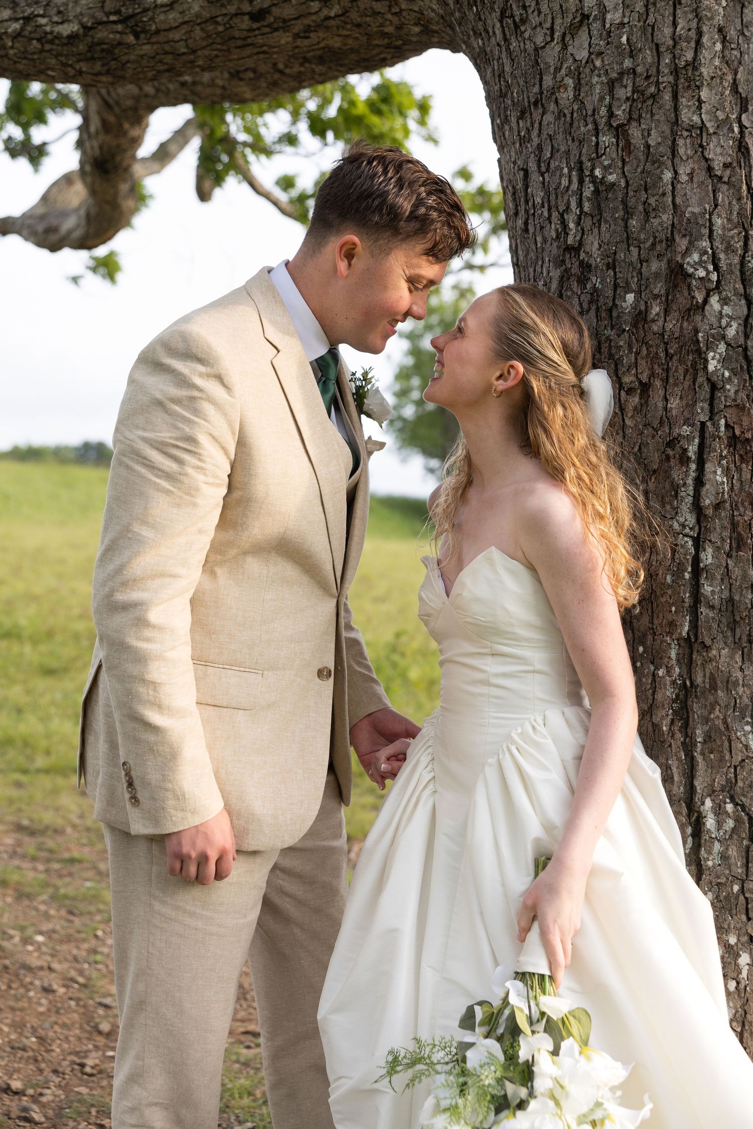Bride and groom share a romantic moment leaning against a tree, bride holding white bouquet in outdoor wedding setting.