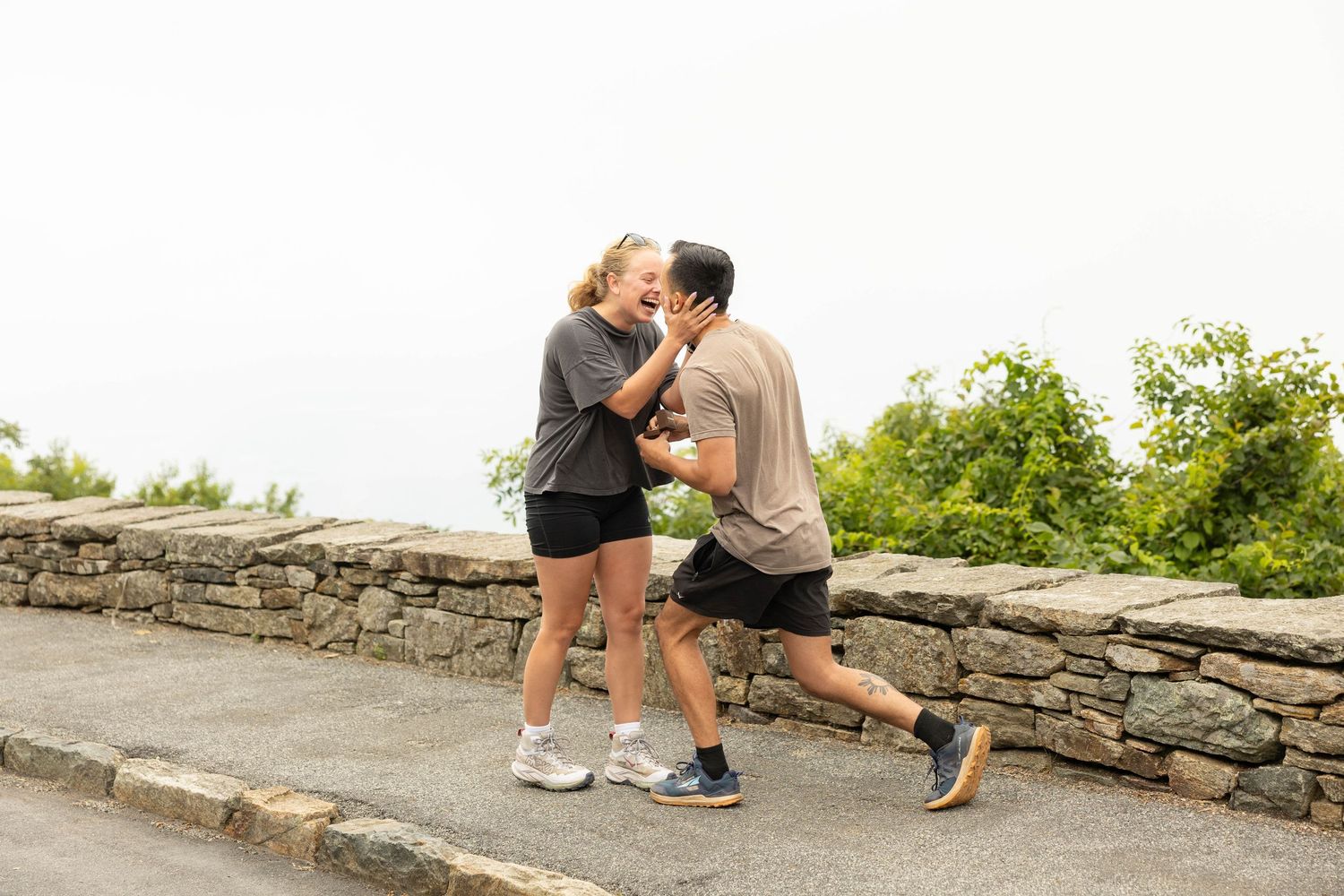 Couple sharing a romantic moment on a stone wall overlook surrounded by lush greenery on a cloudy day.