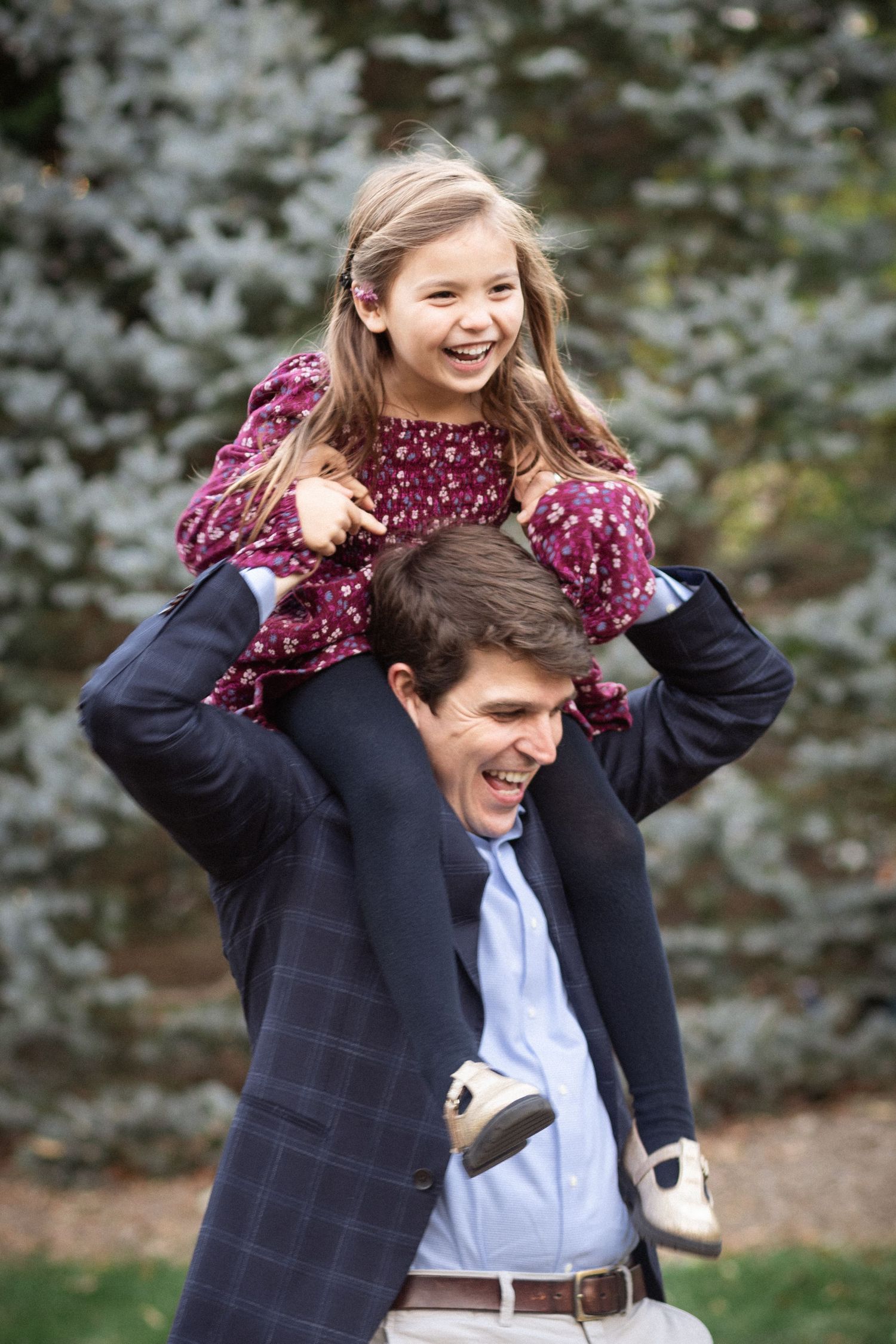 A laughing girl in a floral dress sits on a man's shoulders outdoors near evergreen trees.