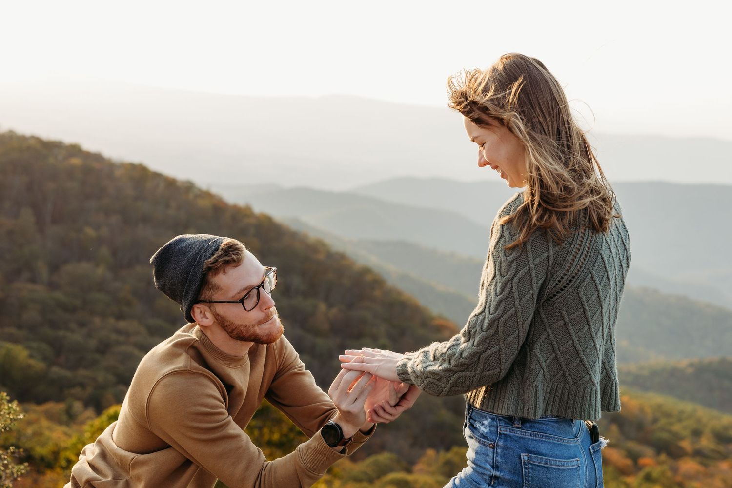 Man kneeling on mountain overlook proposing to woman with scenic autumn valley views in background.
