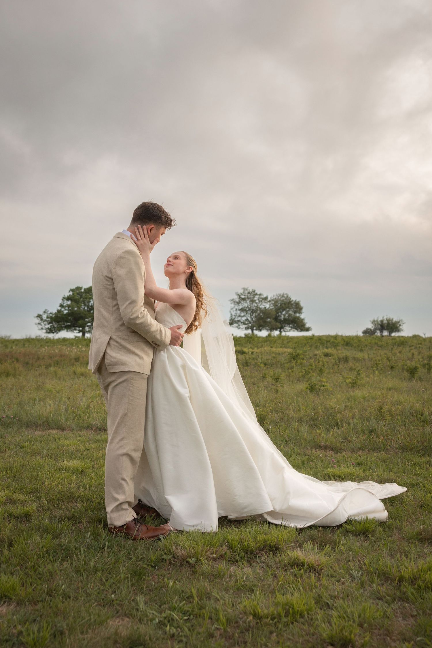 Bride and groom embrace in a green field wearing white gown and tan suit under a cloudy sky.