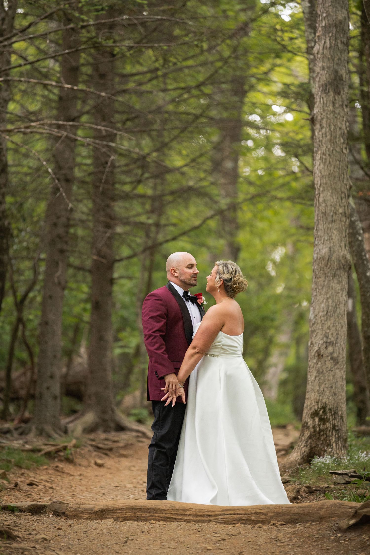 Bride and groom hold hands on a forest path, she in white gown, he in burgundy tuxedo, surrounded by tall trees.