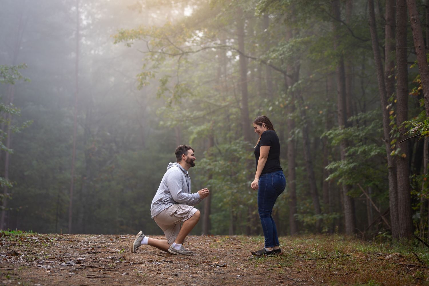 Man kneeling on one knee proposing to woman on a misty forest path surrounded by tall green trees.