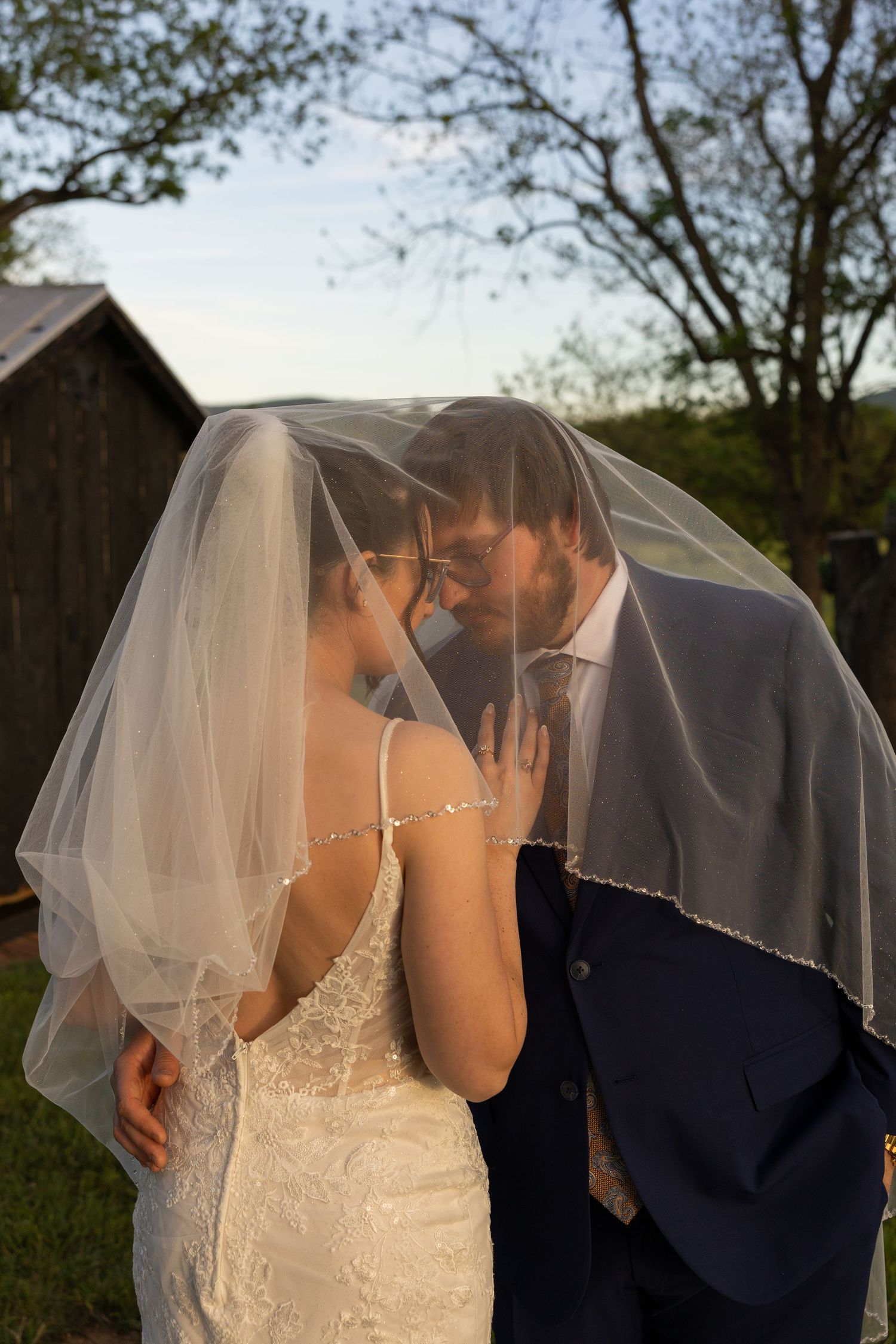 Bride and groom sharing a romantic moment under a flowing wedding veil outdoors near a rustic barn.
