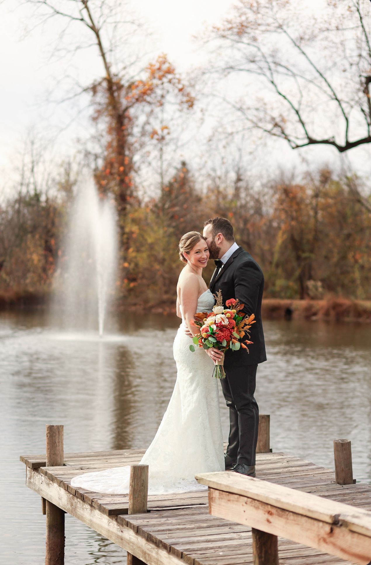 A couple shares an intimate moment on a wooden dock overlooking a lake with a fountain in autumn.