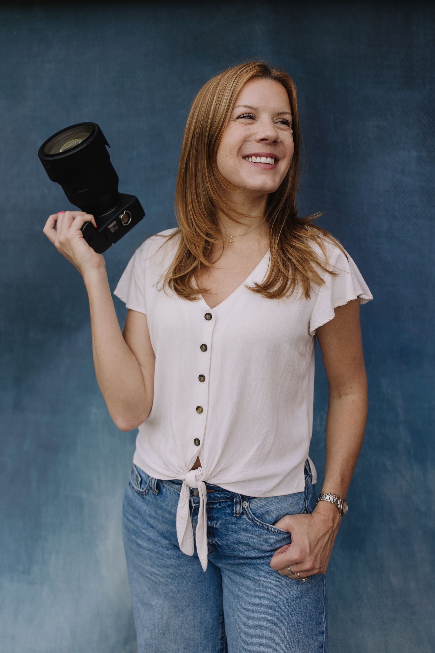 Smiling woman with auburn hair holds a camera lens, wearing a pink blouse and jeans against a blue backdrop.
