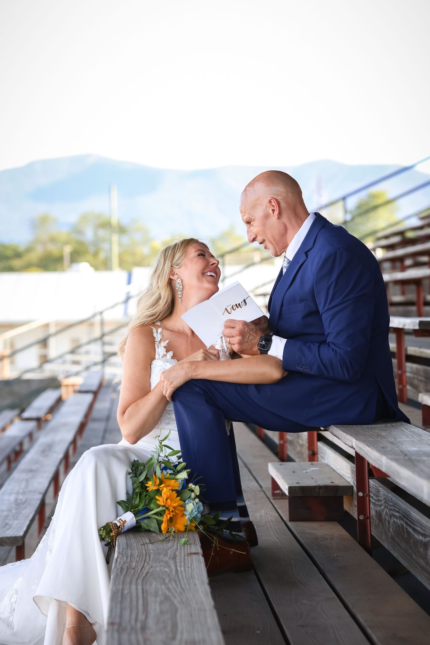 Bride and groom laughing together on bleachers with mountain backdrop, bride holding bouquet of yellow flowers.