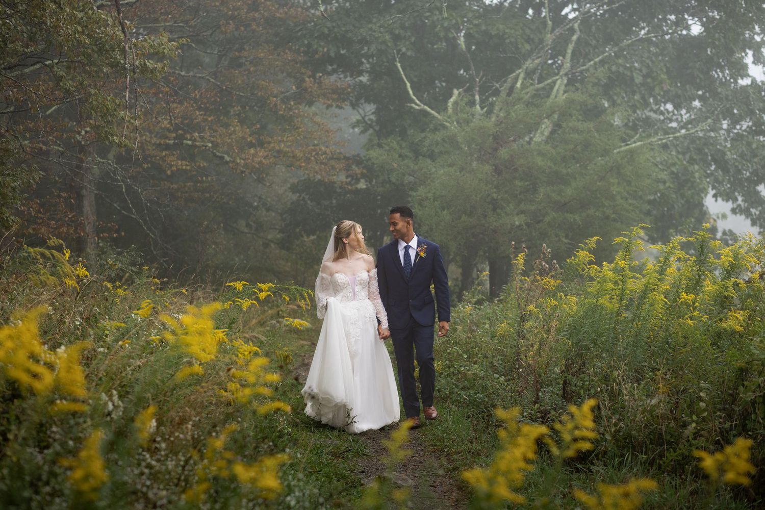 Bride in a white gown and groom in a navy suit hold hands on a misty forest path lined with yellow wildflowers.