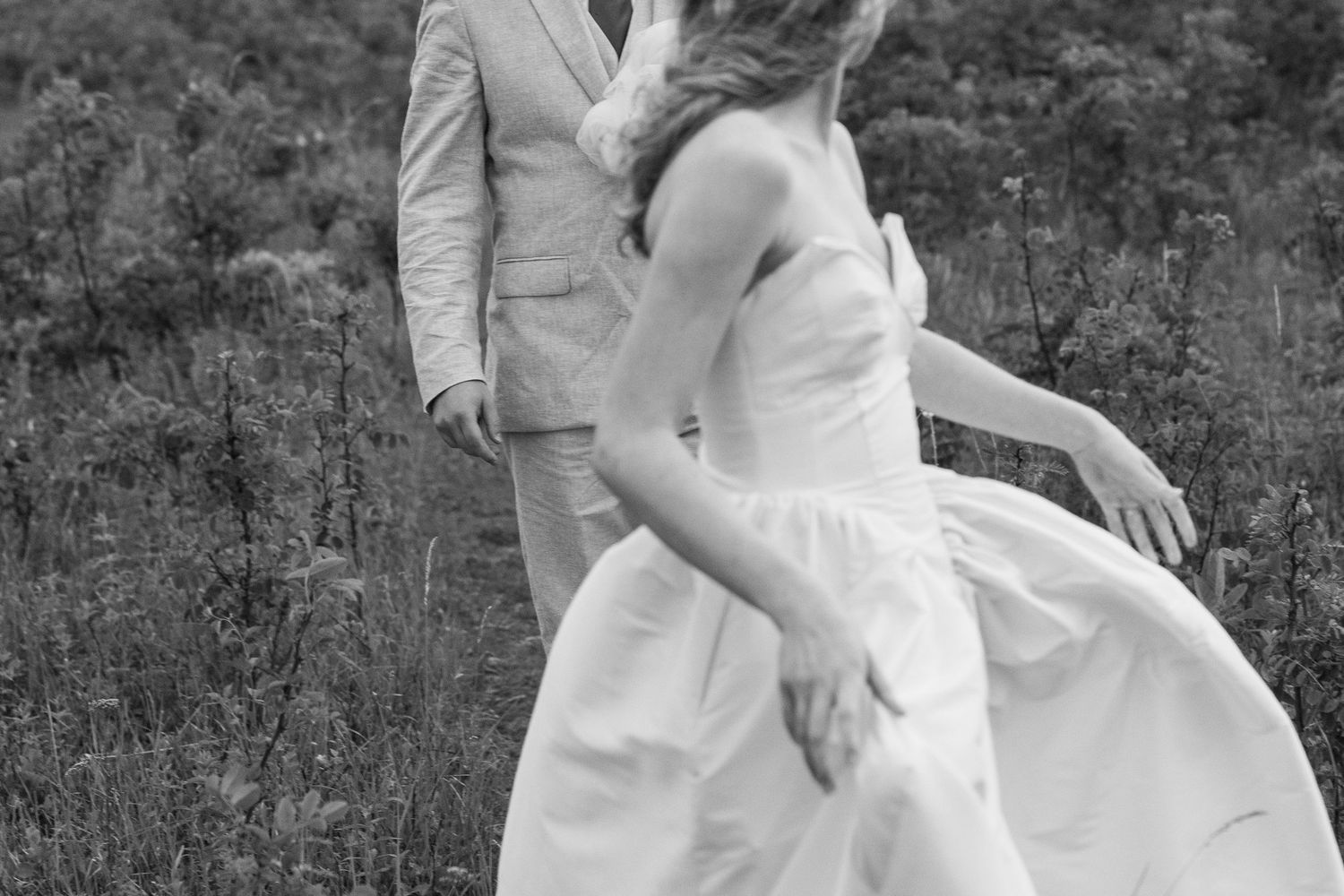 Black and white photo of a bride in a white gown and groom in a suit walking through a field.