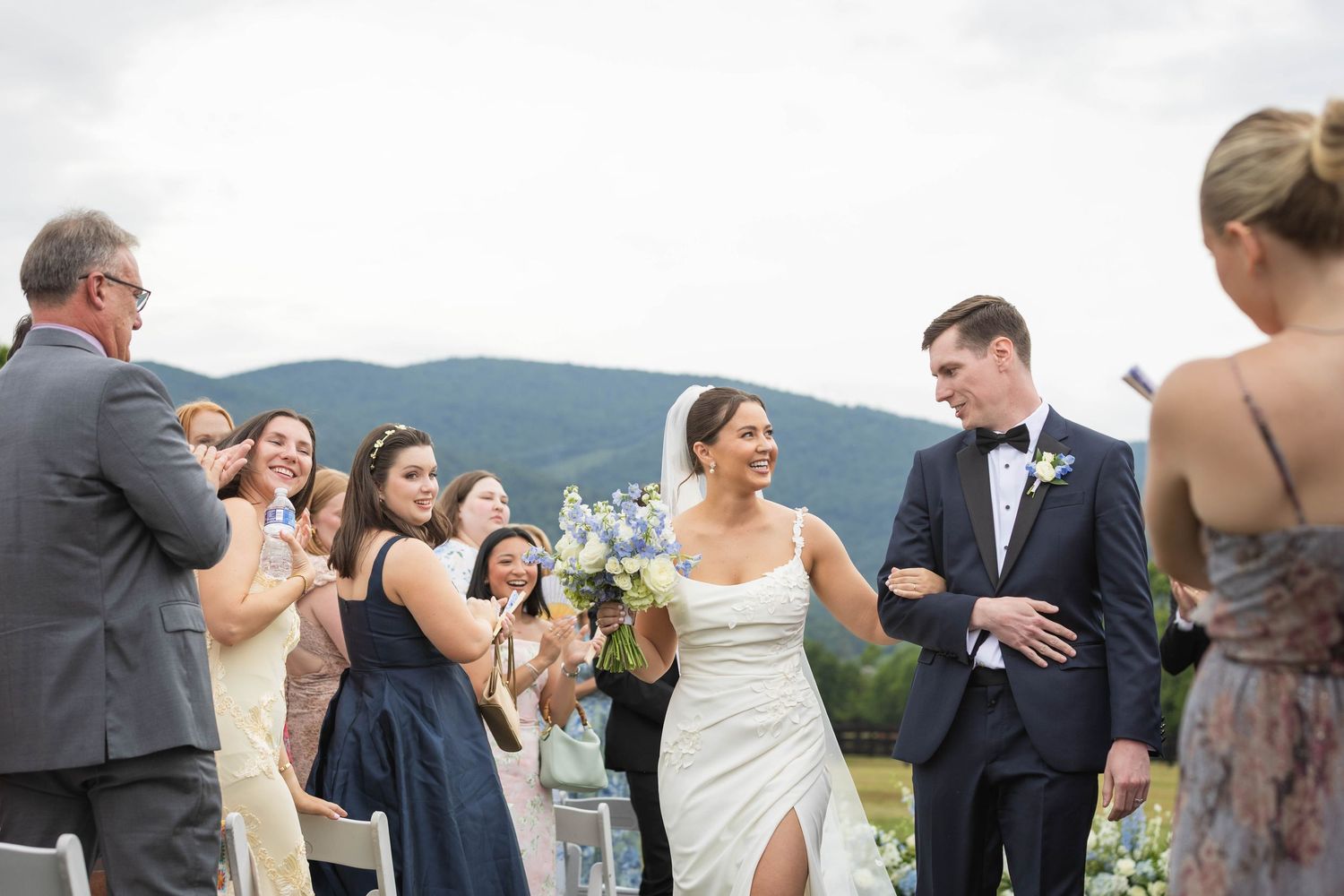 Bride and groom walk down the aisle smiling, surrounded by cheering guests, with mountain views in the background.