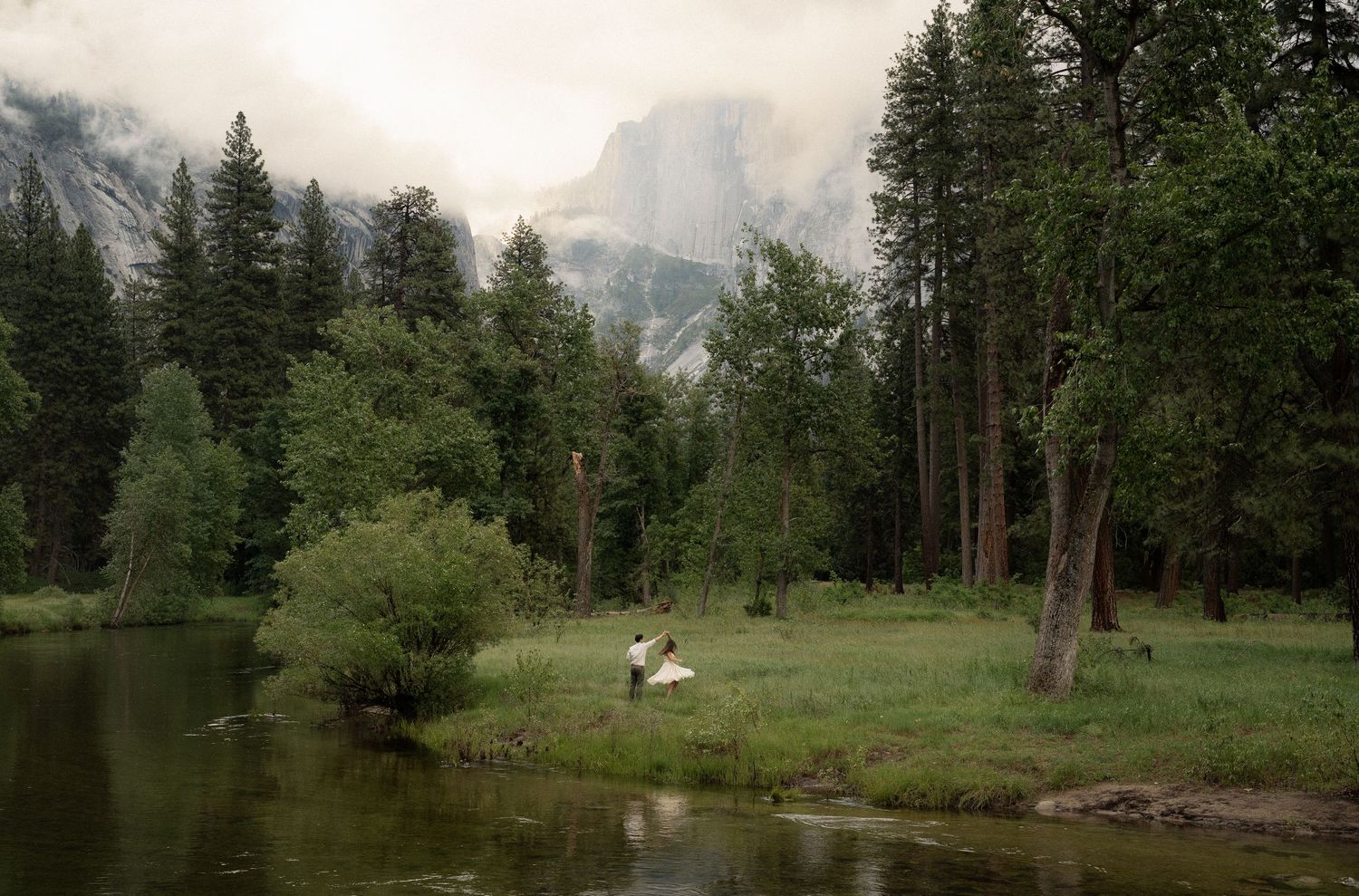 Couple in wedding attire by a serene river in Yosemite Valley with lush trees and misty mountains in the background.