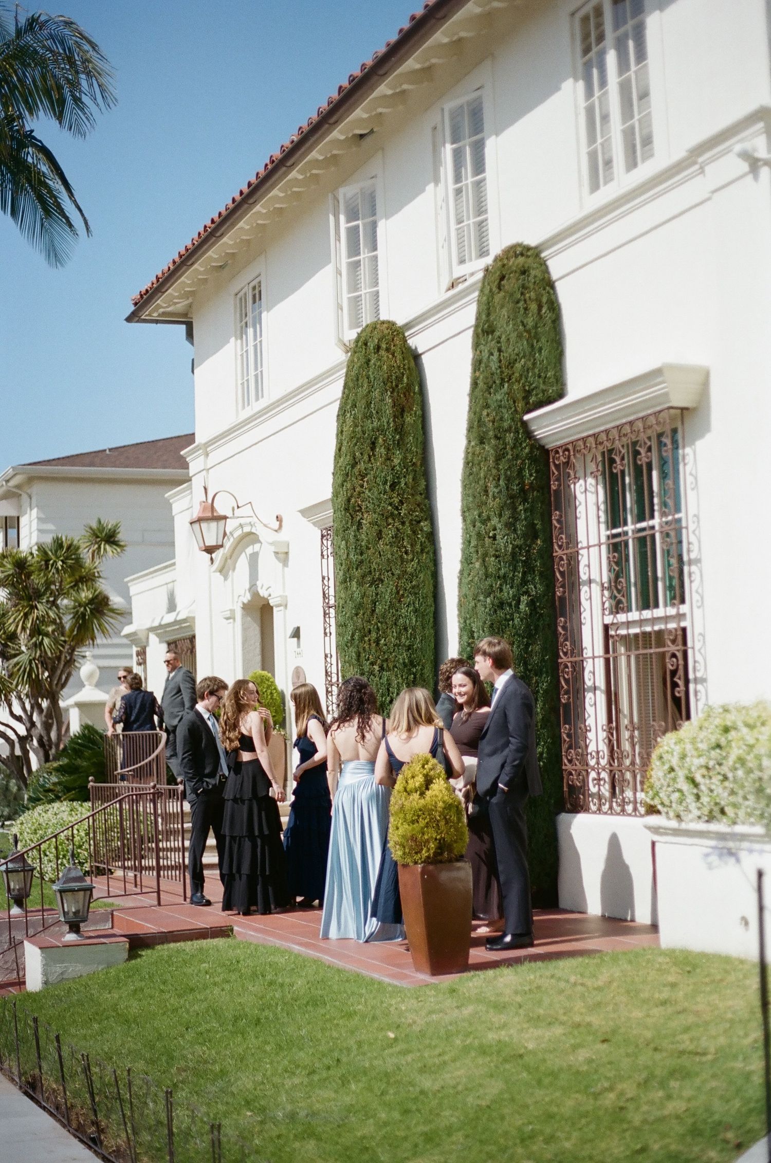 Elegantly dressed guests mingle outside a white Mediterranean-style villa with cypress trees on a sunny day.