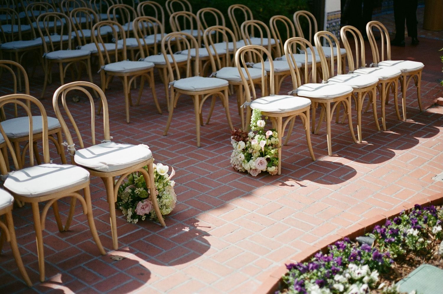 Rows of wooden bentwood chairs with white cushions arranged for an outdoor wedding ceremony on a brick patio with floral arrangements.