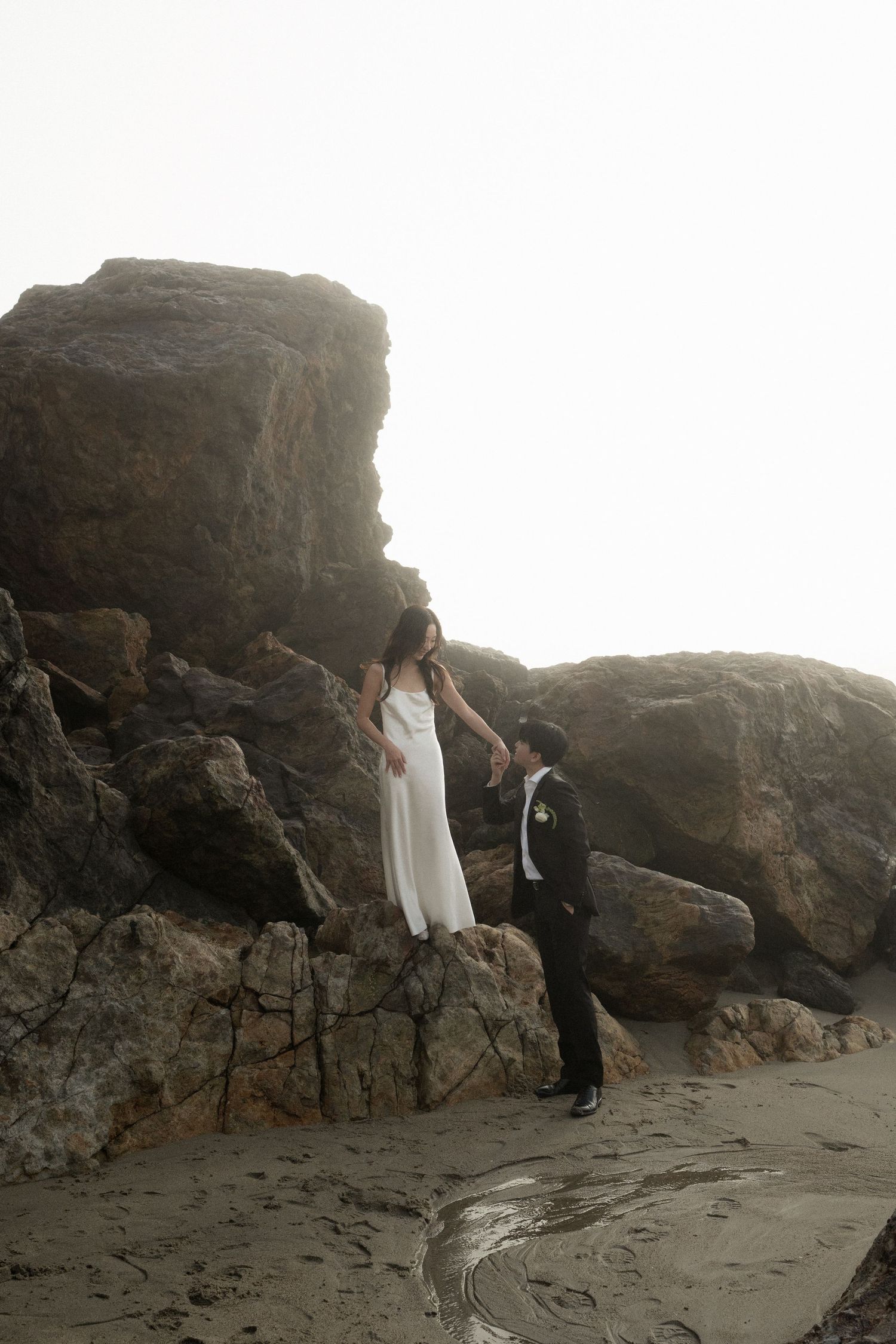 A couple in formal attire poses romantically on a misty beach with dramatic rocky cliffs in the background.