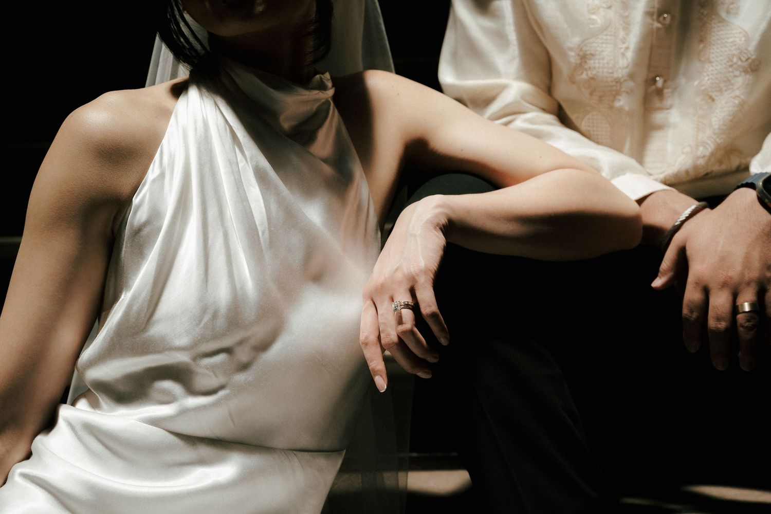 Two people seated closely together with intertwined hands, one wearing a white sleeveless top and the other in dark clothing.