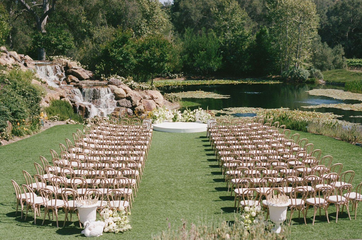 Outdoor wedding ceremony setup with rows of wooden chairs on green lawn, floral altar, pond and waterfall backdrop.