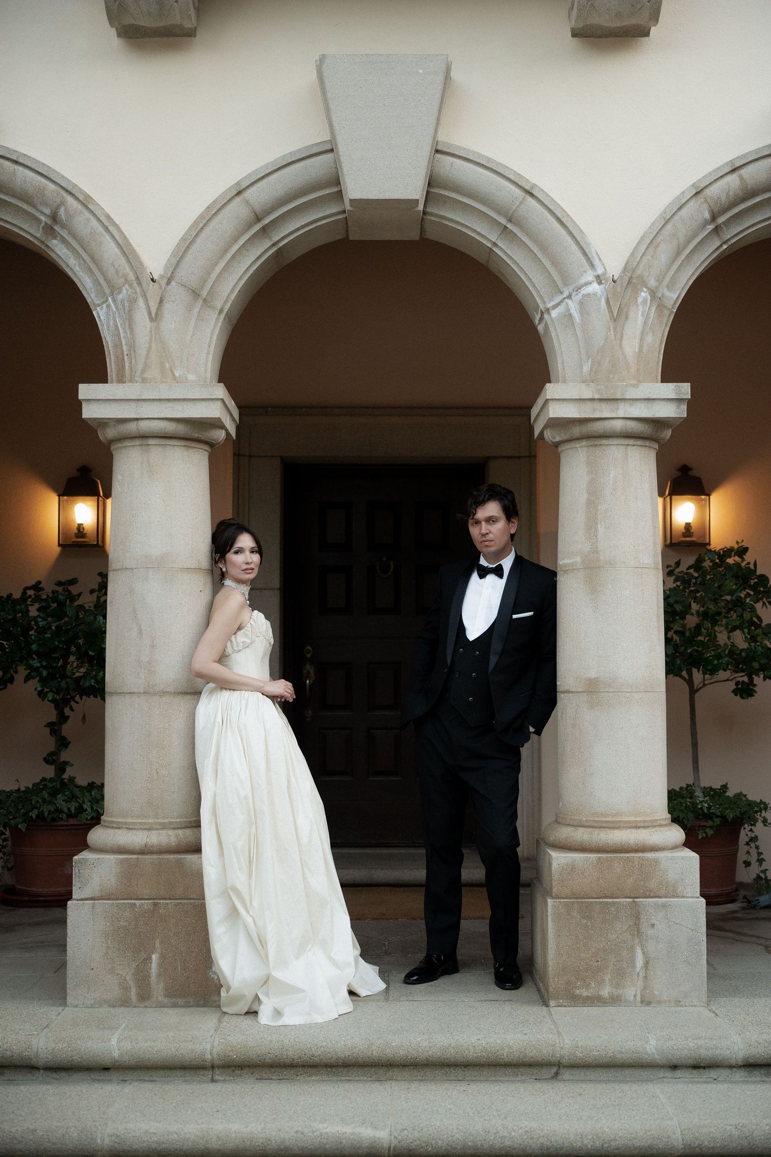 A bride in a white gown and groom in a black tuxedo stand between elegant stone archways with warm lighting.