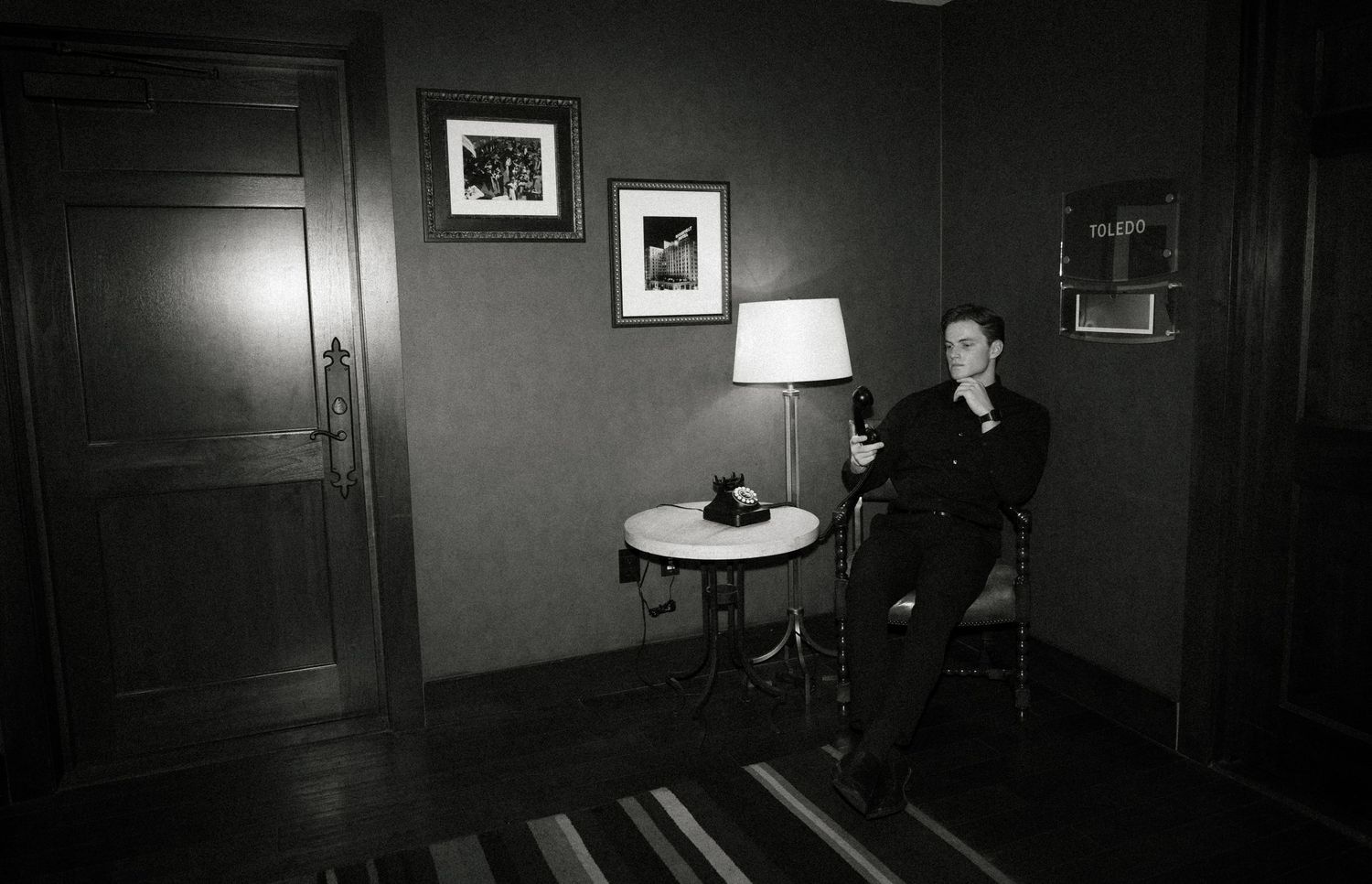 A vintage-style black and white photo of someone sitting in a dimly lit room with a table lamp.