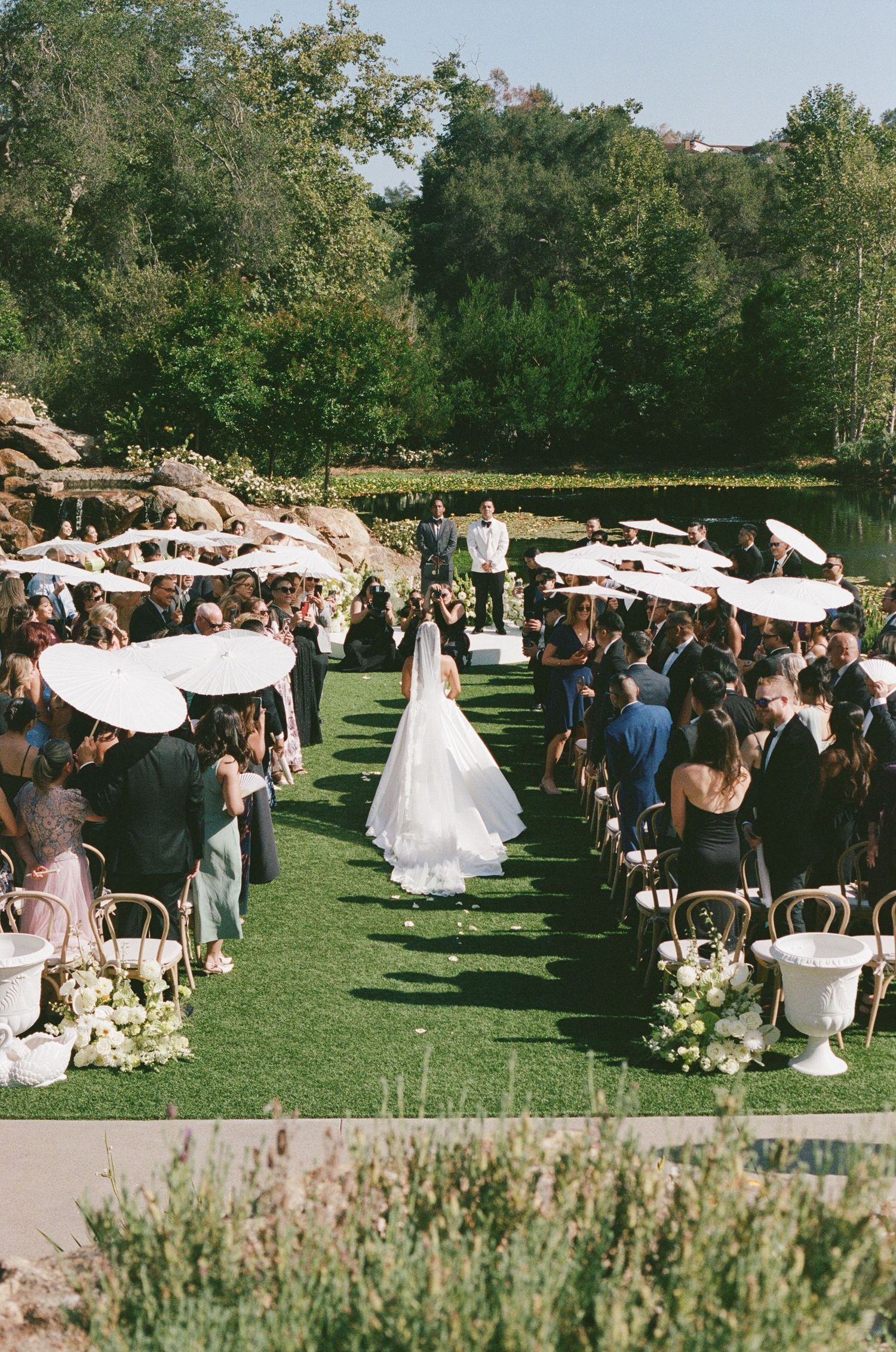Bride walks down outdoor grass aisle lined with white parasols and floral urns toward altar with lush greenery backdrop.