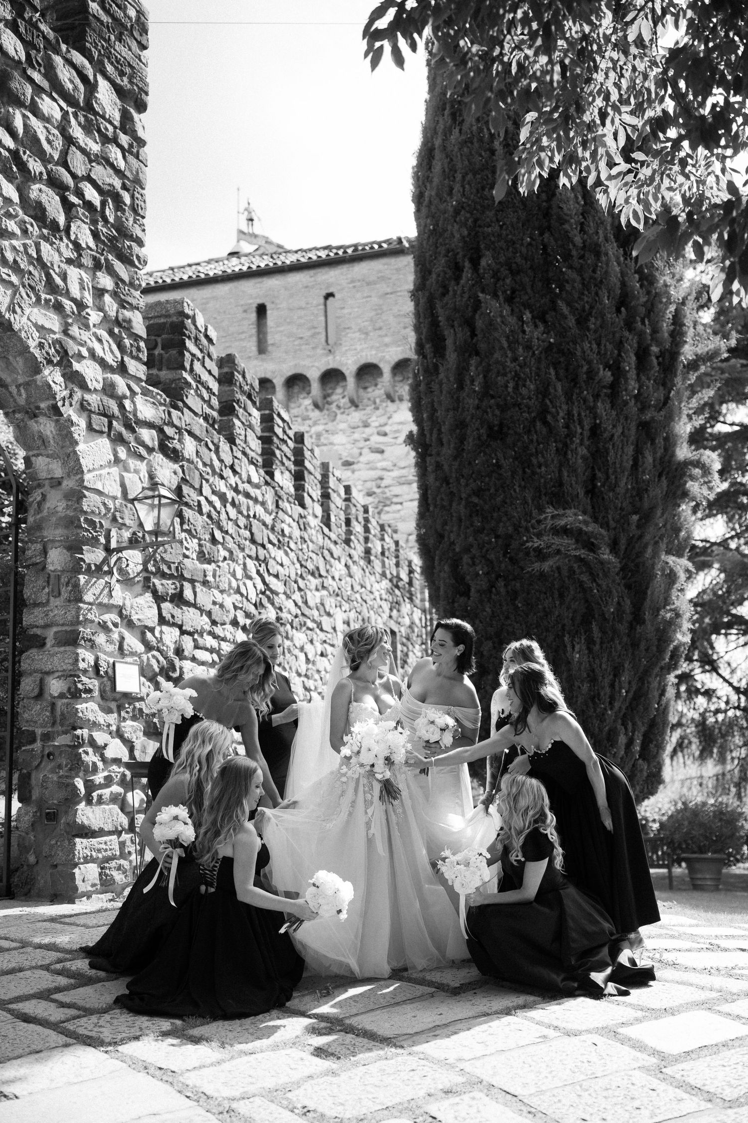 A wedding party in black and white poses against a stone castle wall with cypress trees in an elegant outdoor setting.