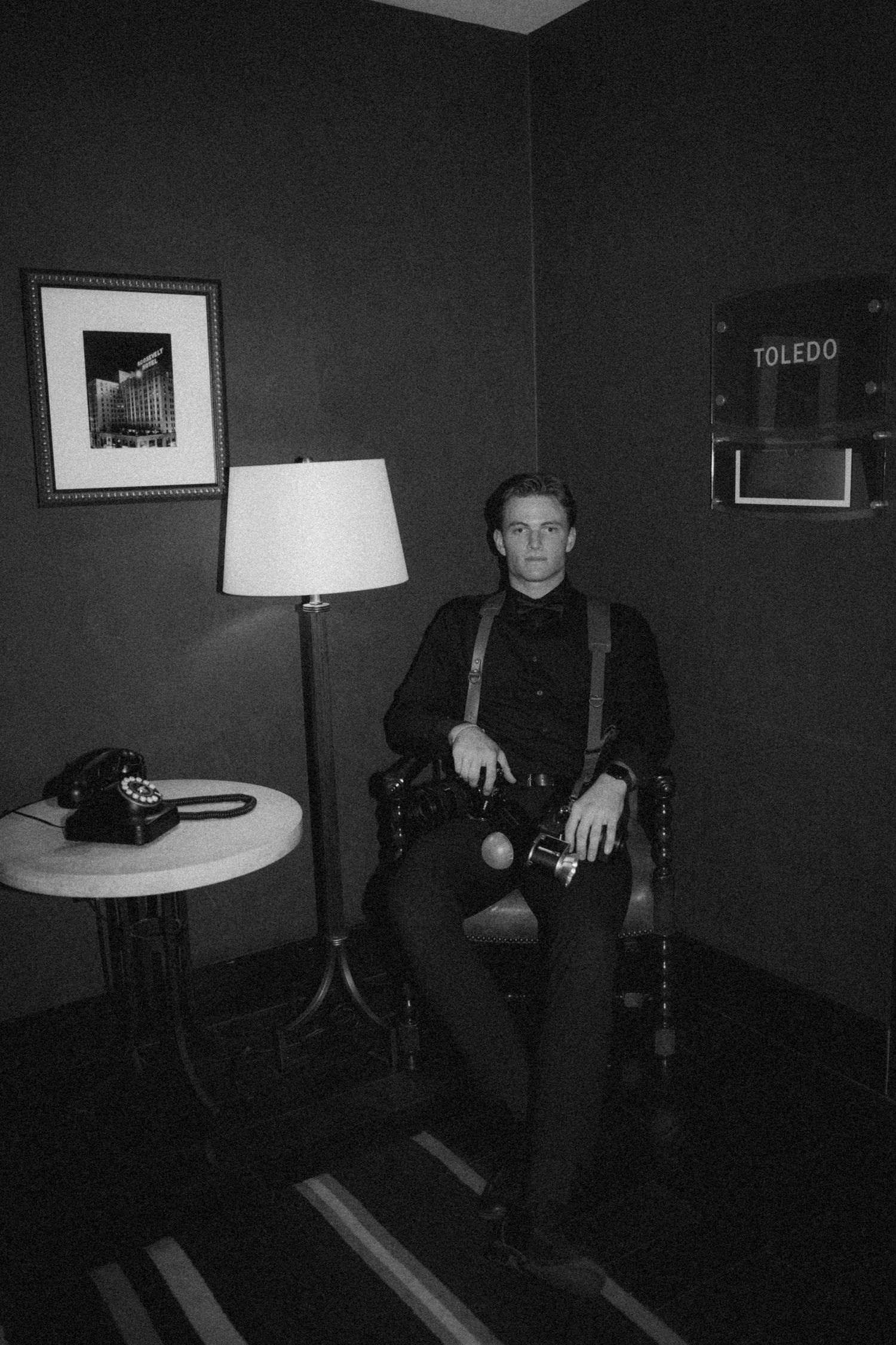 A dramatic black and white portrait of a person in suspenders seated in a vintage-style room with a lamp.