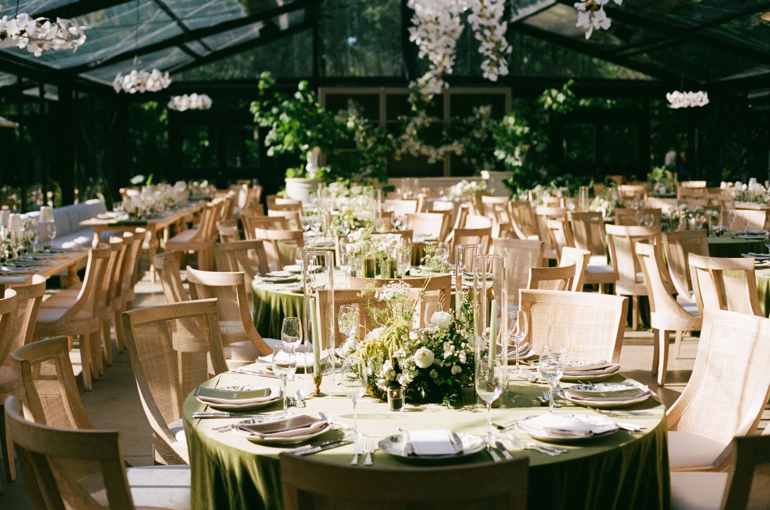 Elegant wedding reception in a greenhouse venue with round tables draped in green linen and floral centerpieces.