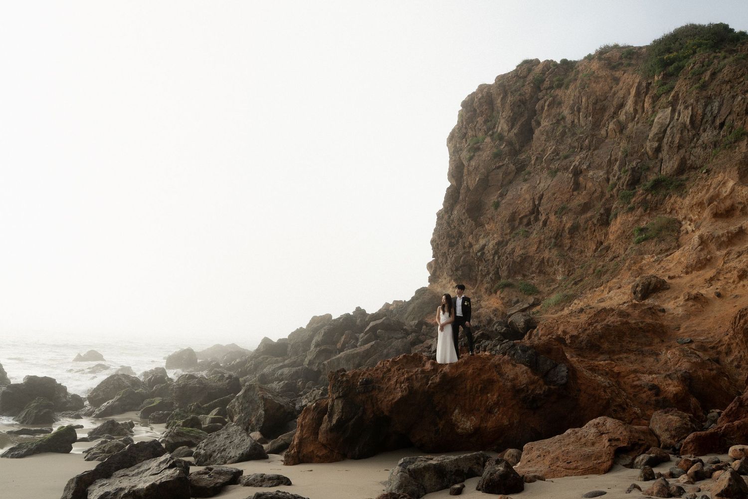 Panoramic view of rocky coastal cliffs and misty ocean waves crashing against the rugged California shoreline at sunset.