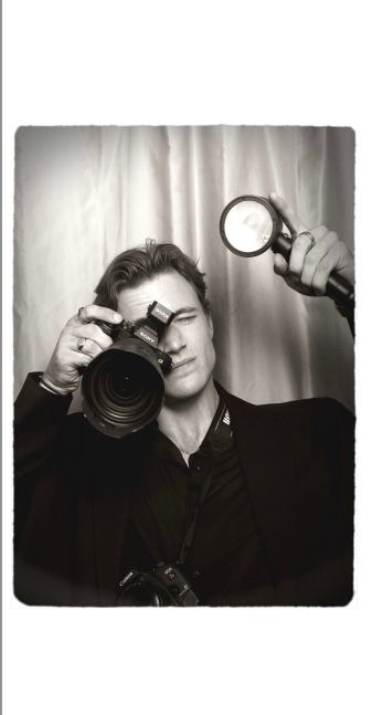 Black and white photo of a man holding a Sony camera and flash, dressed in black, posing as a professional photographer.