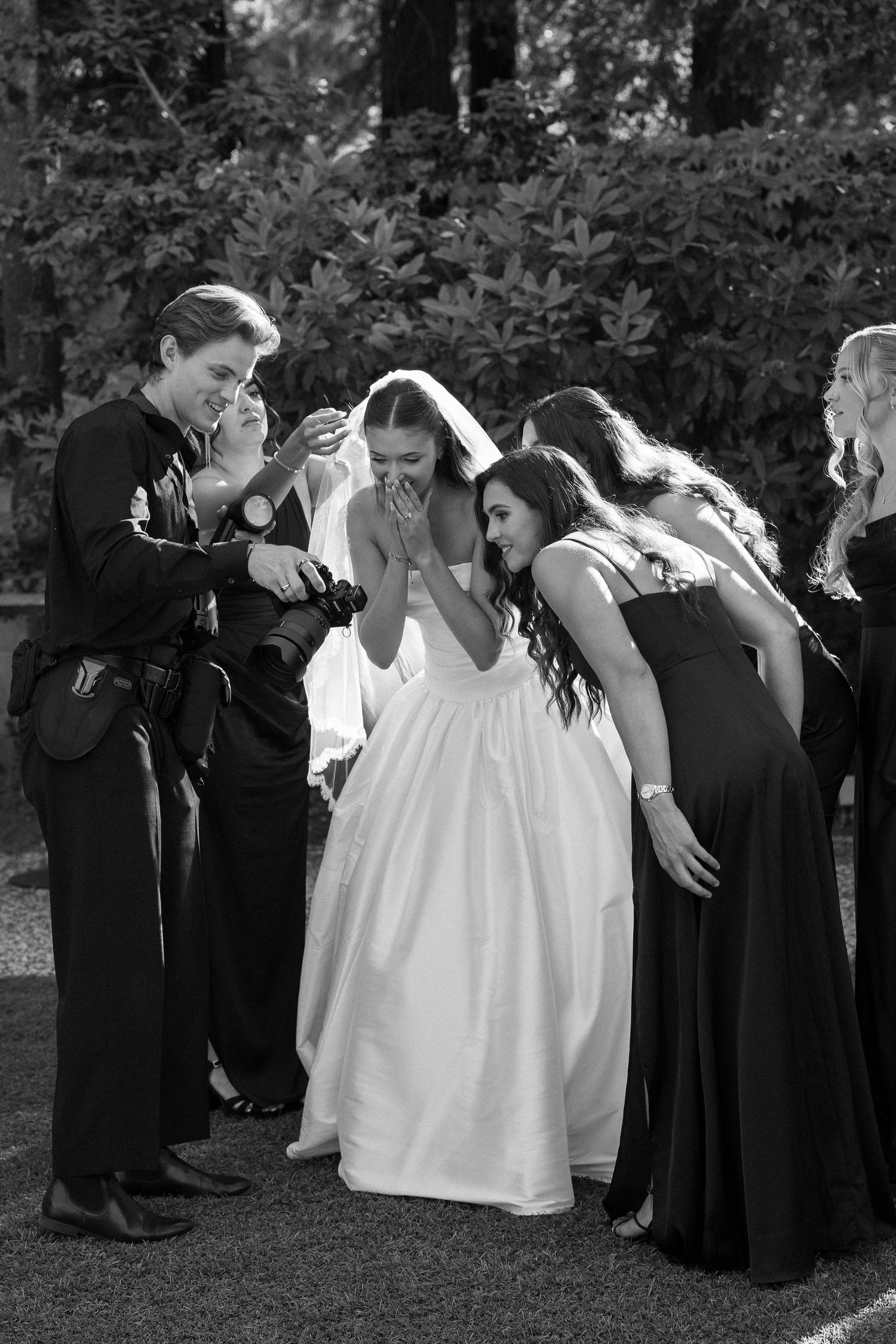 Black and white photo of a bride in white gown and veil laughing with bridesmaids in dark dresses at outdoor wedding.