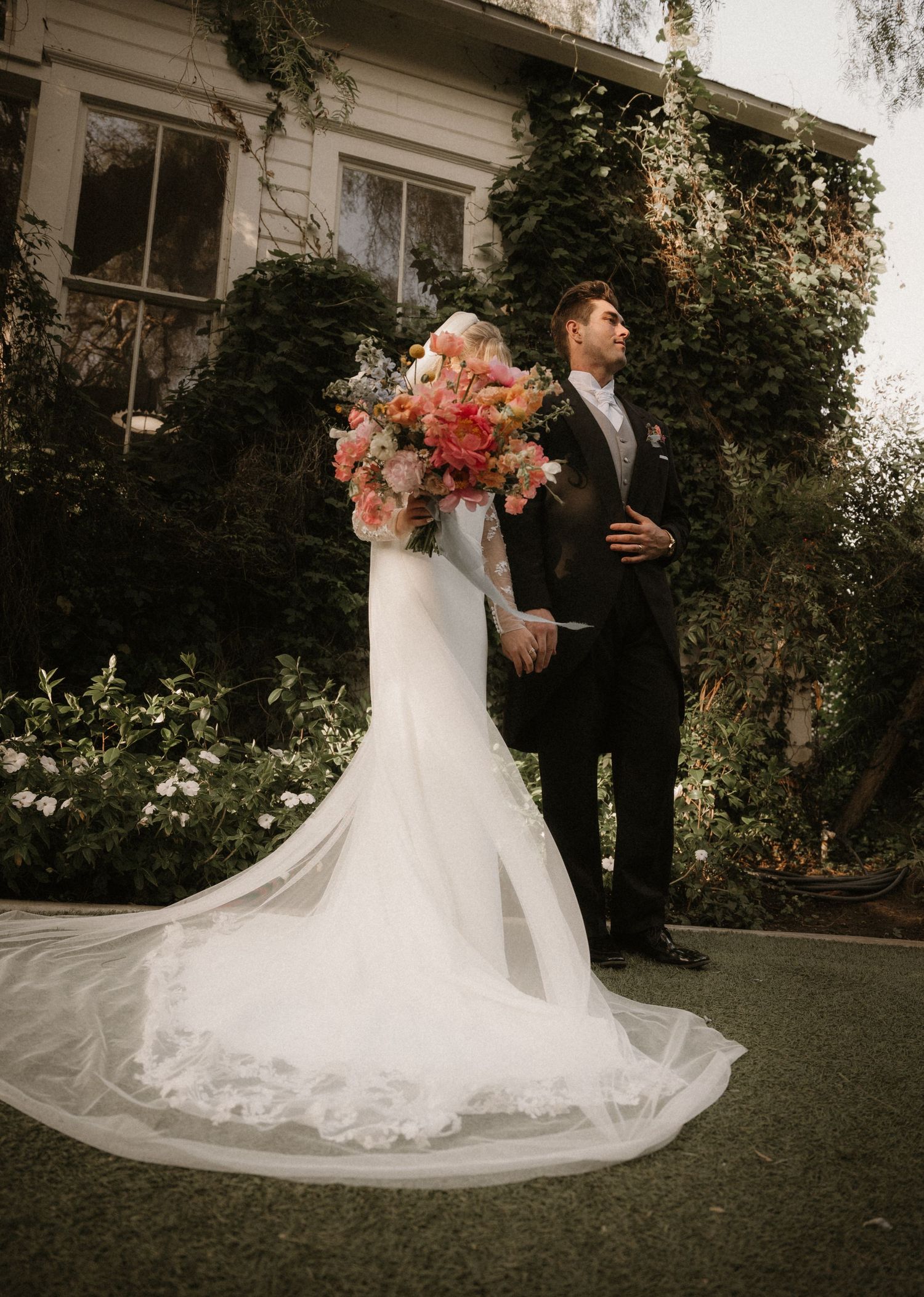 A classic wedding photo showcasing a stunning white mermaid gown and large coral bouquet against an ivy-covered backdrop.