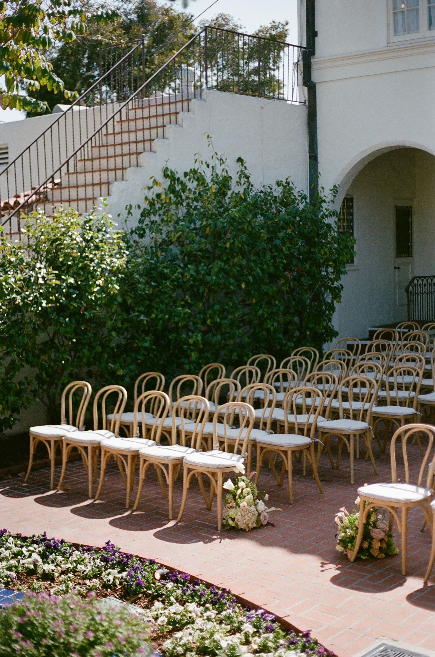 Outdoor wedding ceremony setup with bentwood chairs arranged in rows on a brick patio beside a white villa with lush greenery.