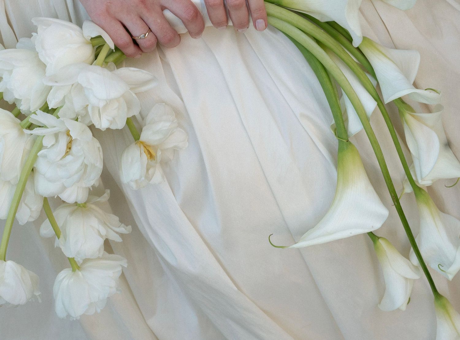 Elegant white calla lilies and tulips arranged on soft ivory fabric with a hand wearing a delicate ring.