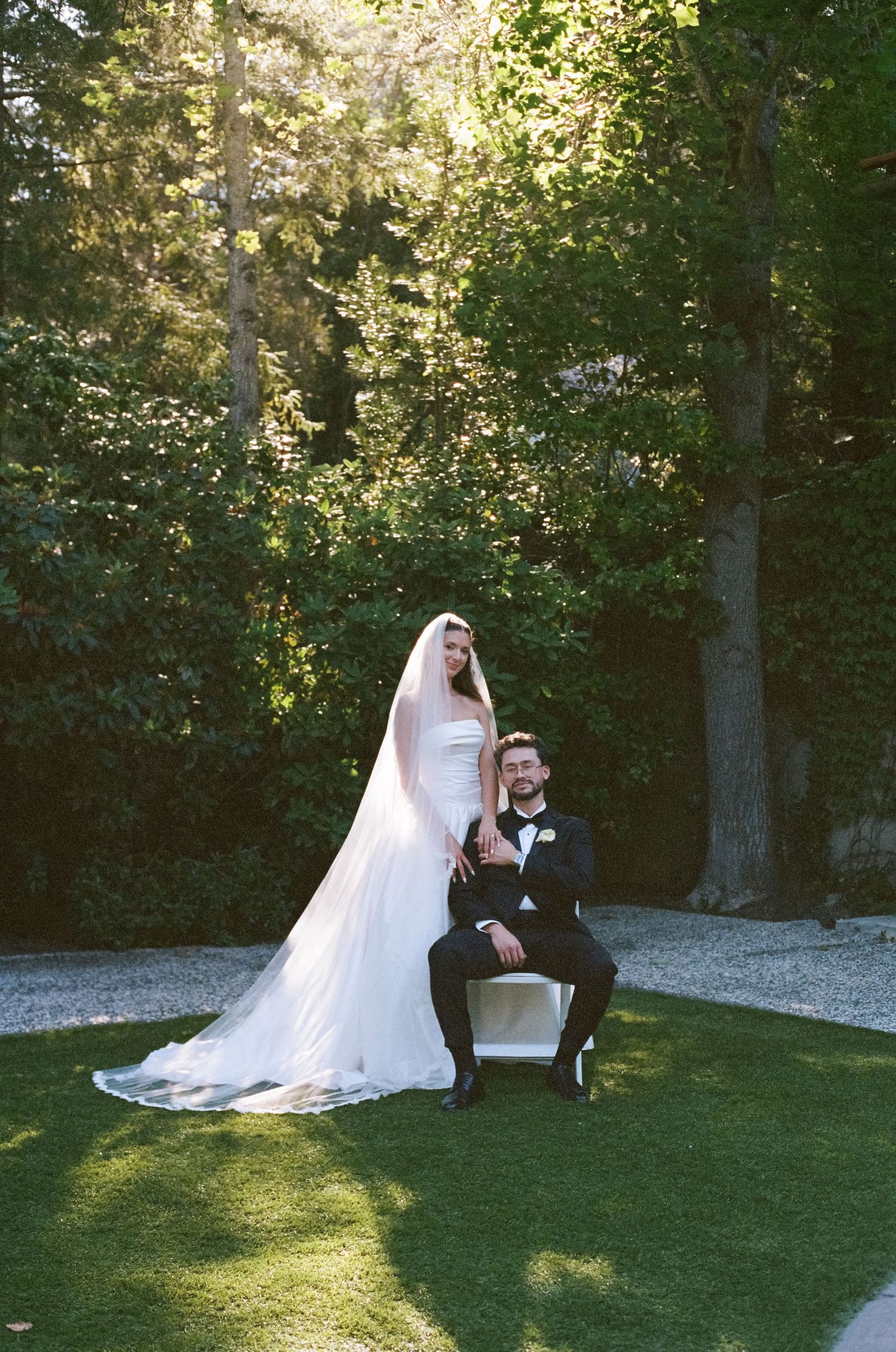 Bride in white gown and veil stands beside groom in black tuxedo seated on chair in lush garden setting.