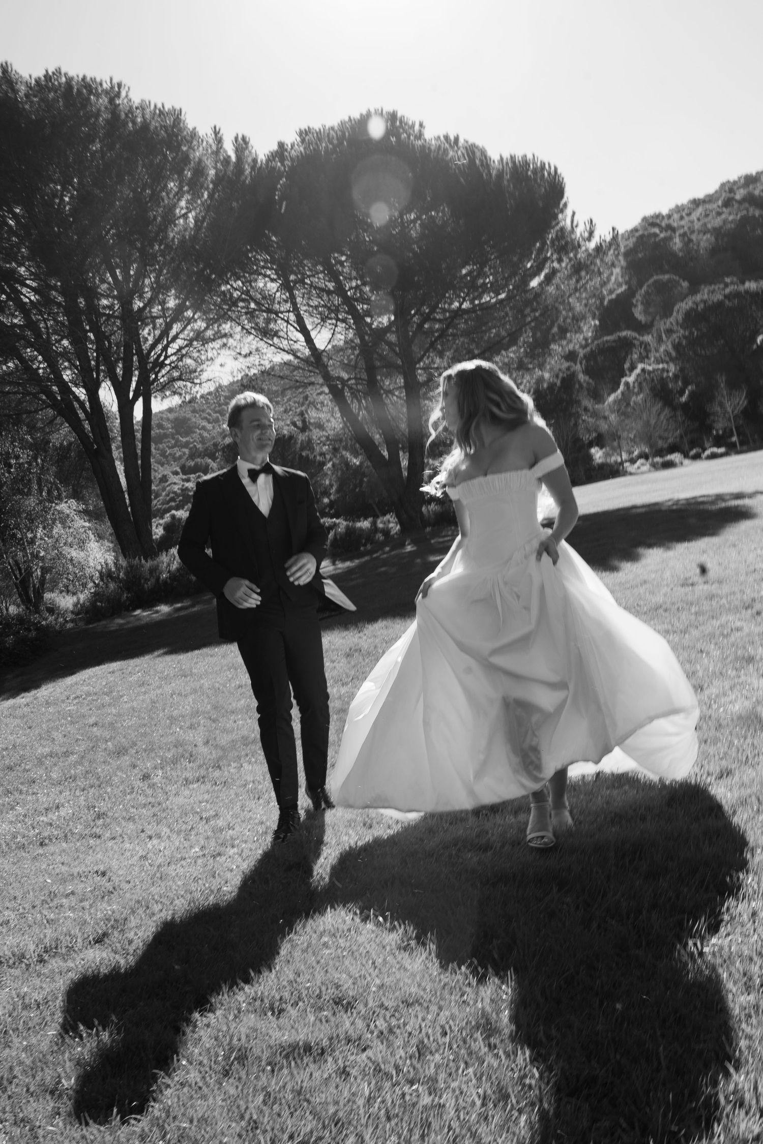 A bride and groom twirl playfully on a grassy lawn in black and white, creating romantic silhouettes against the trees.