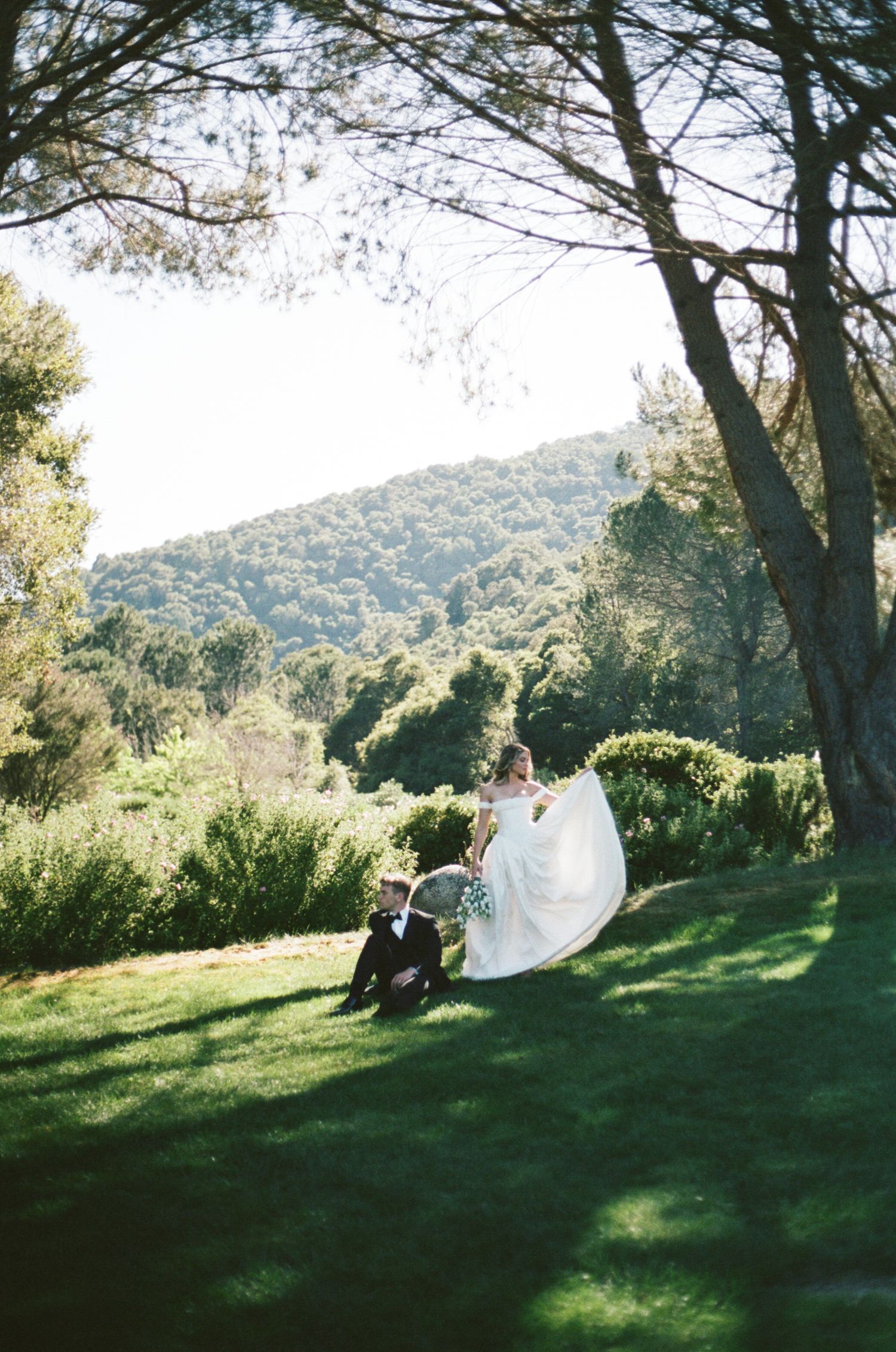 Bride in white gown twirls on lush green lawn as groom sits nearby, with scenic forested hills in background.