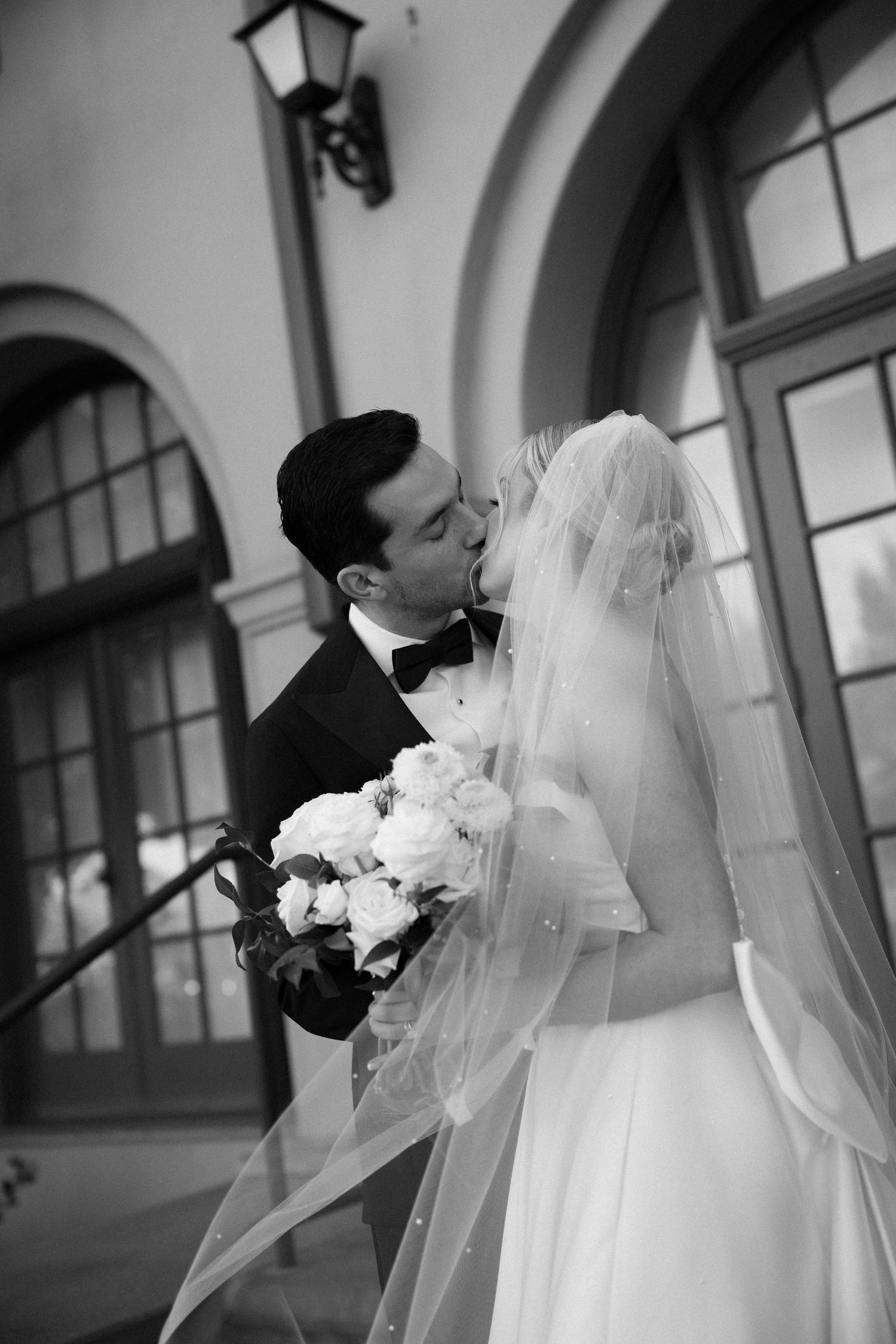 A romantic black and white wedding photograph showing a tender kiss between newlyweds outside an arched doorway.