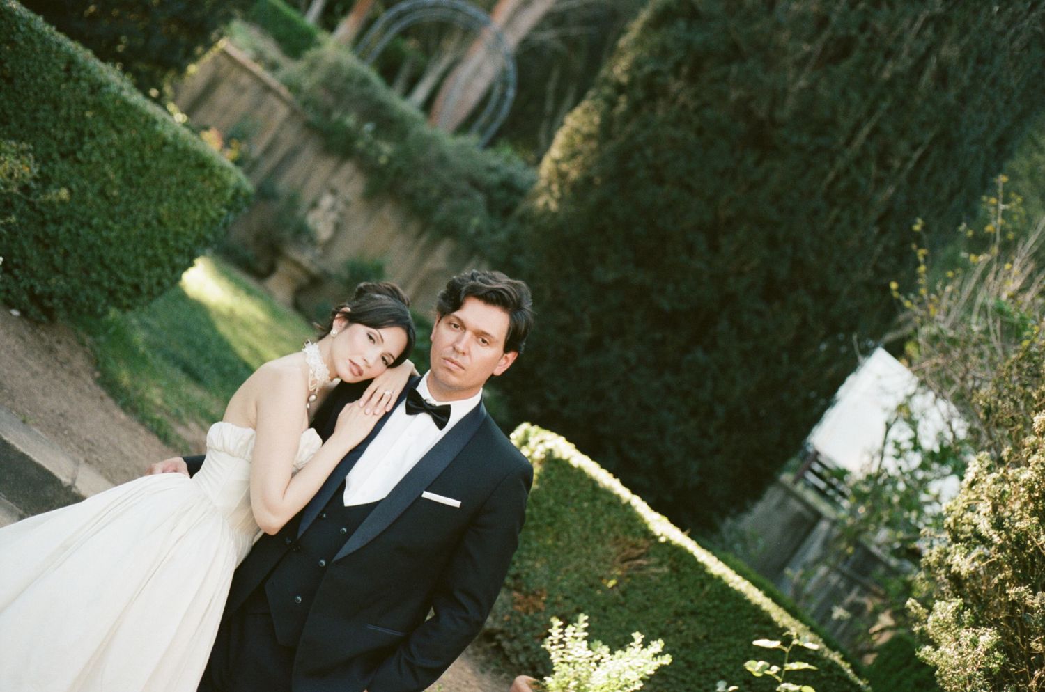 A couple in formal wedding attire poses together in a lush garden setting during their outdoor wedding celebration.