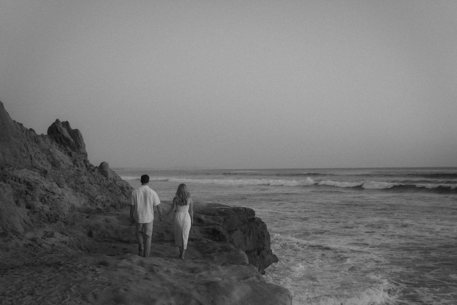 A black and white coastal photograph of two people standing on rocky cliffs by the ocean.
