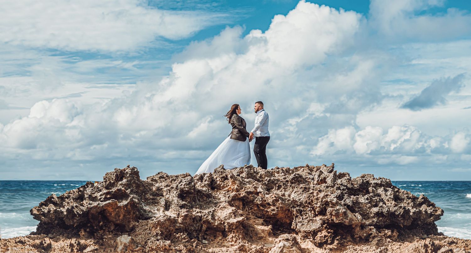 Two people stand atop a rocky cliff overlooking the ocean under a dramatic blue sky with white clouds.